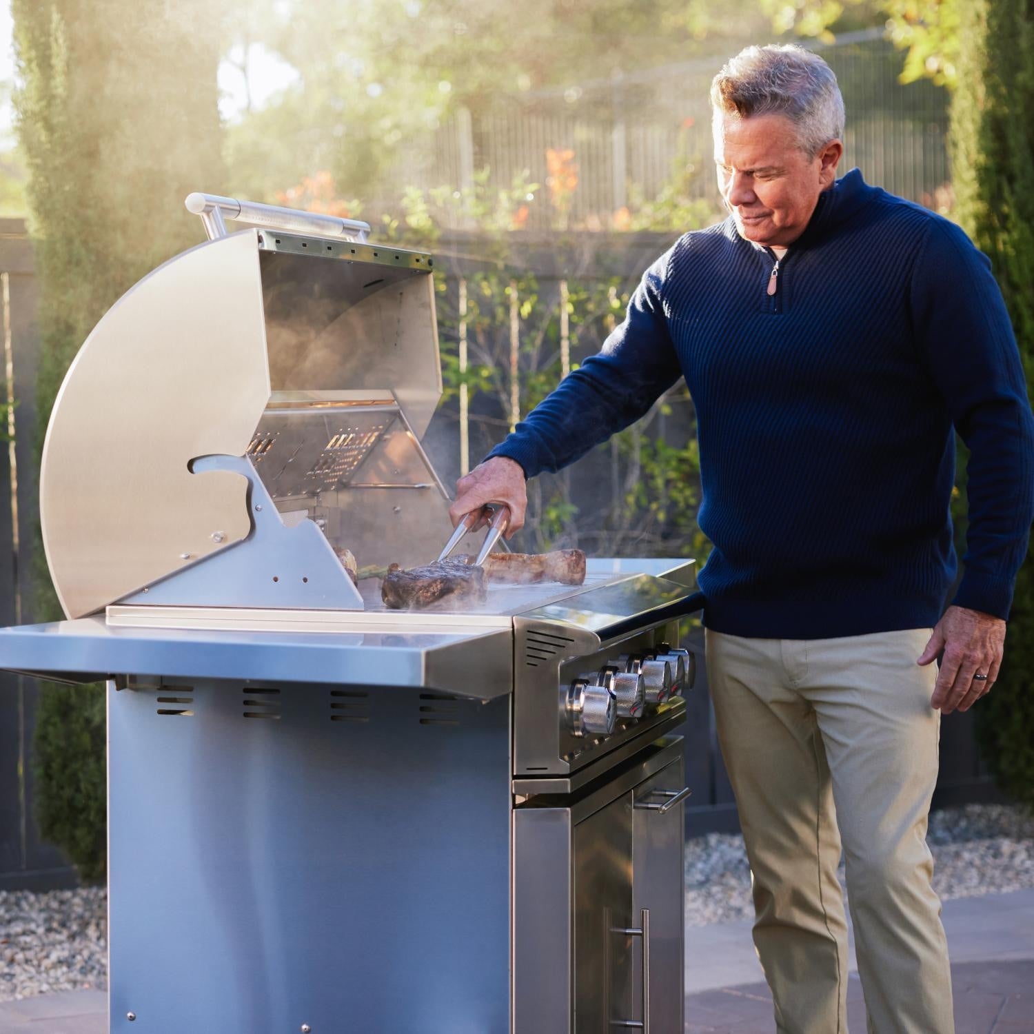 Man grilling outdoors using a large barbecue grill in a garden setting.
