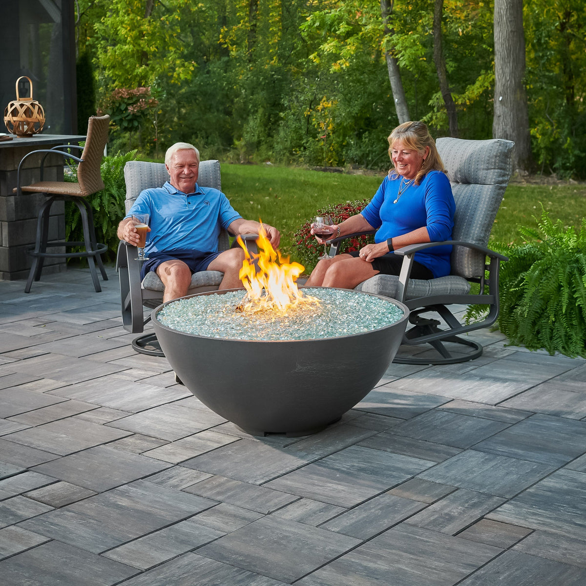 Two people beside a Midnight Mist Cove Concrete Edge 42-inch gas fire pit bowl in a backyard, vertical view