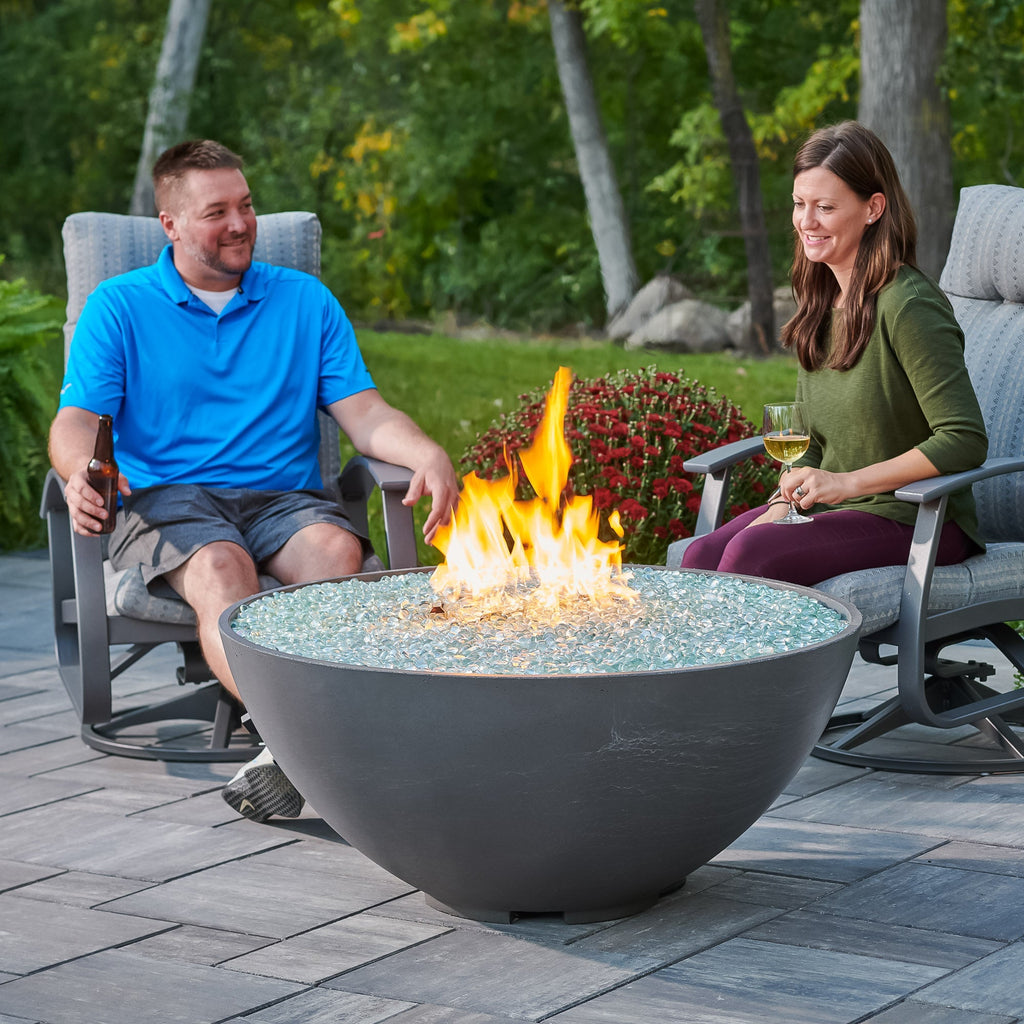 Two people relaxing beside a Midnight Mist Cove Concrete Edge 42-inch gas fire pit bowl on a patio