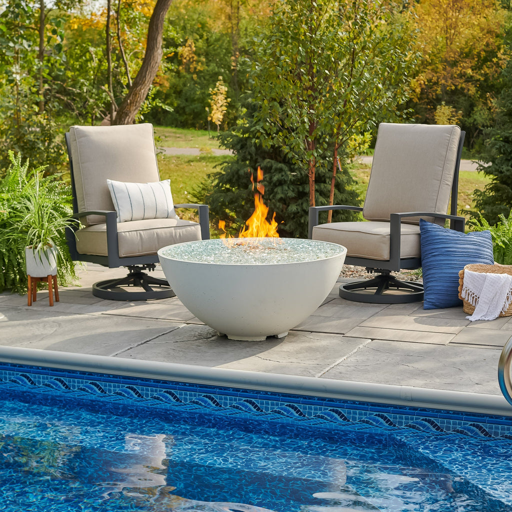 White Cove Concrete Edge 42-inch gas fire pit bowl with lit flame beside a pool and lounge chairs